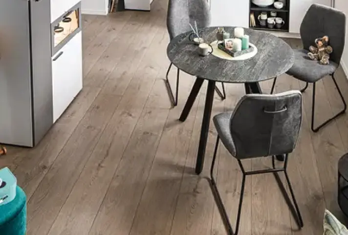 Runder dunkler Esstisch mit drei grauen Polsterstühlen auf Holzfussboden, auf dem Tisch Tablett mit Kerzen, dahinter weisse Küchenmöbel mit offenem Regal und Geschirr.